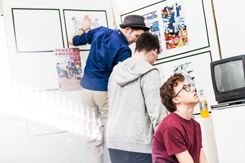 2 Male students hanging artwork on white wall; one male student looking up at another wall of art.