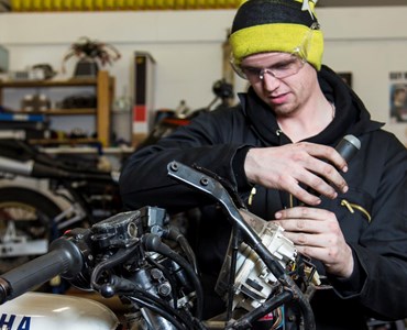 Male student in yellow beanie hat, protective goggles and black overalls working on a section of a motorcycle.