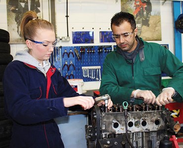 Female and male both in overalls and protective goggles working on an engine.