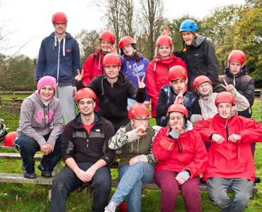 Large group of students sat together smiling in red helmets and harnesses.