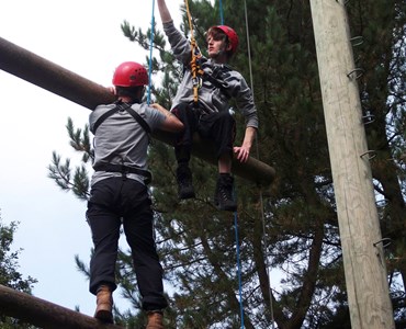 Two students in harnesses and red helmets climbing a Jacob's ladder.