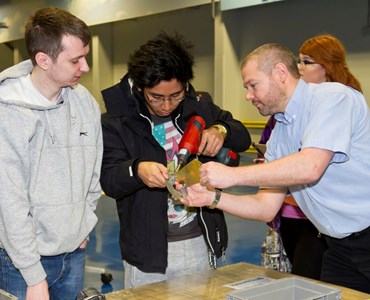 Lecturer stood with two students helping them use a power drill.