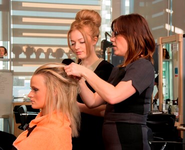 Blonde  female sat in hairdressers chair with orange towel around her shoulders; female lecturing styling hair and speaking with female student who is observing.