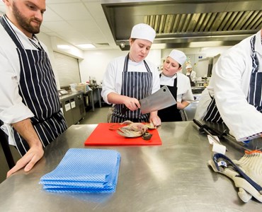 Hospitality students in chef whites and aprons in professional kitchen; student with large knife and meat on chopping board.