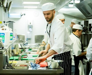 Male in chef whites and apron preparing food in a hospitality kitchen.