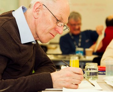 Mature male student painting with water colours at a desk in a classroom environment.