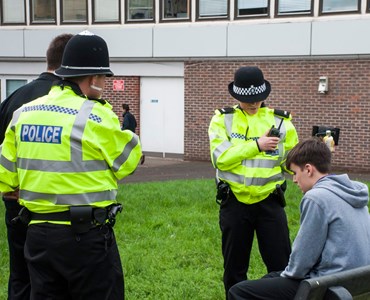 2 Police officers in high vis jackets, talking to a young male sat on a bench with another male stood next to one of the police officers