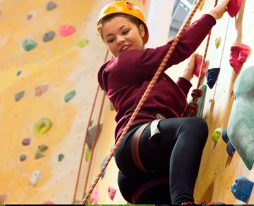 A female student climbing an indoor climbing wall wearing a helmet and harness looking down towards the floor