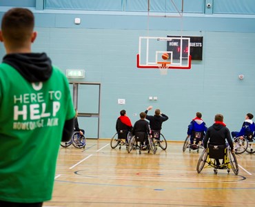 A group of male students playing indoor wheelchair basketball with a referee in a green t-shirt stood watching