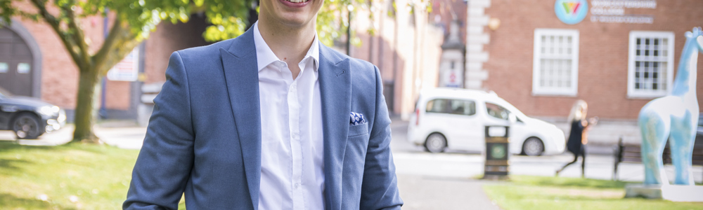 Male former student wearing a grey-blue suit stood outside of HoW College building