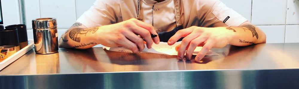 Male wearing a chefs uniform leaning on stainless steel shelf and writing on a piece of paper