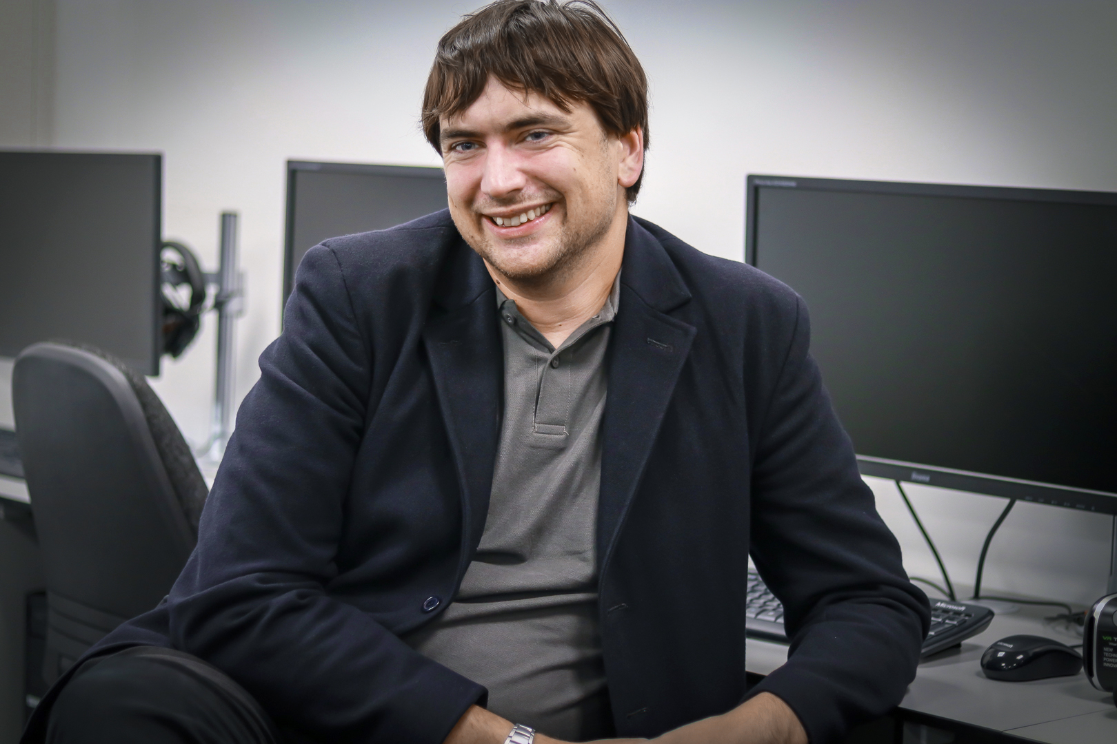 Male in Navy blazer smiling and sat in front of computer screens