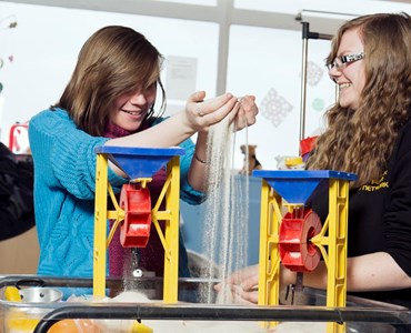 Two female students playing in sand box and laughing.
