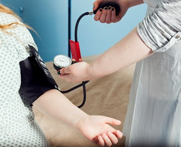 Female using blood pressure machine to measure blood pressure of female sat down in hospital gown.