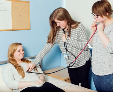 Female sat on medical bed while two female students monitor heart beat with dual stethoscope 