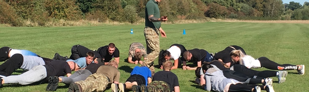 Group of male and female students holding the plank position in a large circle in the middle of a field, with a marine holding a stop watch in the middle.