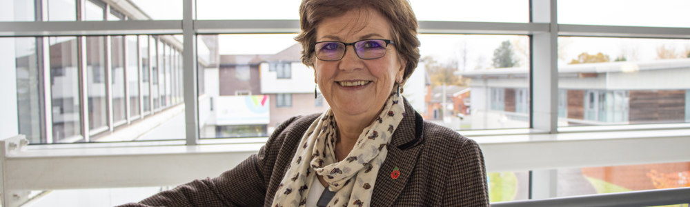 Woman wearing glasses and a cream coloured scarf stood smiling whilst leaning on a railing in front of a window