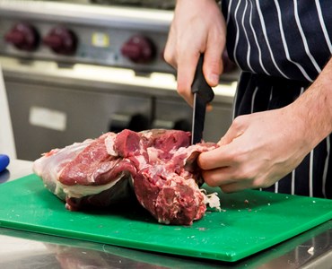 A chef's hand slicing a large piece of red meat with a knife on green chopping board
