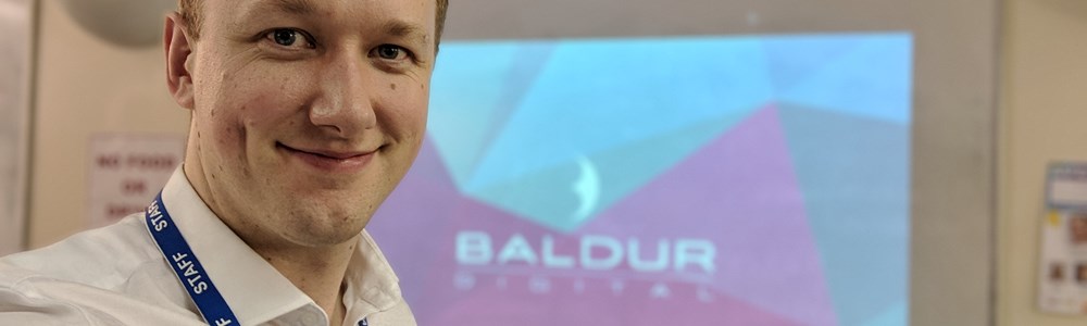 Smiling staff member in a white shirt stood in front of a college whiteboard. Baldur digital logo projected onto said whiteboard.