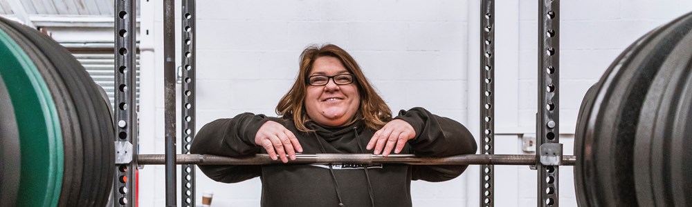 Image of female in black hoodie smiling and resting her arms on a squat bar in a gym
