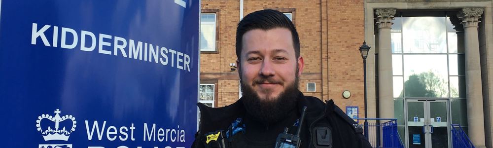 Male police officer with beard stood in uniform and smiling in front of the West Mercia Police Kidderminster sign