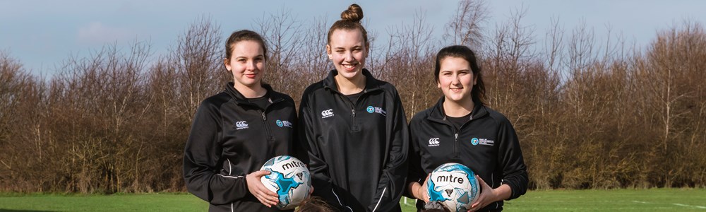 Group of 5 females in matching black football jackets smiling and stood in a field holding footballs.