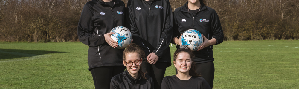 Group of 5 females in matching black football jackets smiling and stood in a field holding footballs.