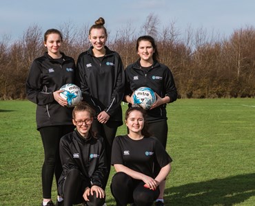 Group of 5 females in matching black football jackets smiling and stood in a field holding footballs.