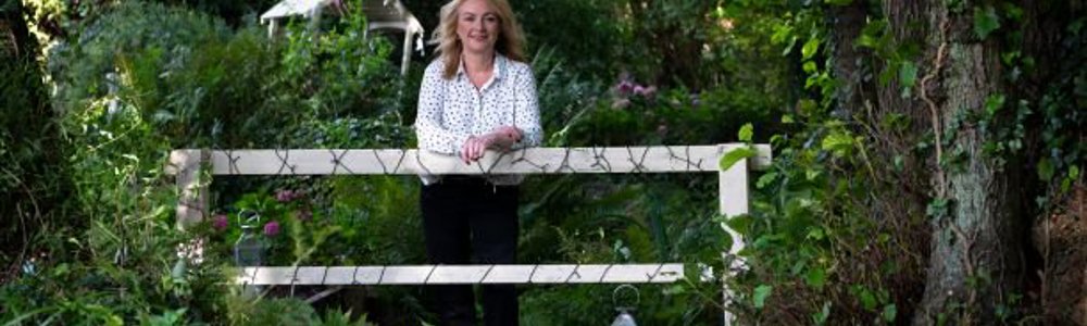 Woman from a distance smiling and leaning on white fence with trees and shrubbery surrounding