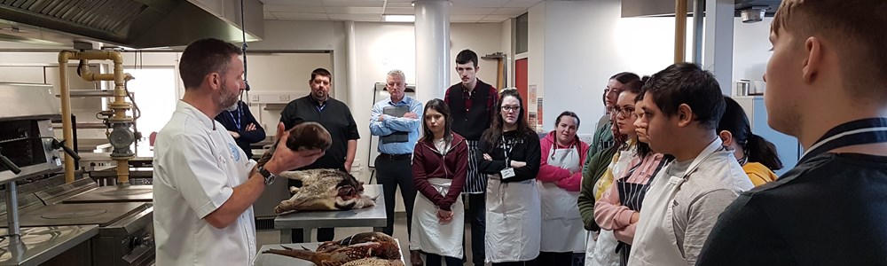 Male lecturer stood behind a stainless steel table in a commercial kitchen. Speaking to a class of students while holding a dead game bird.