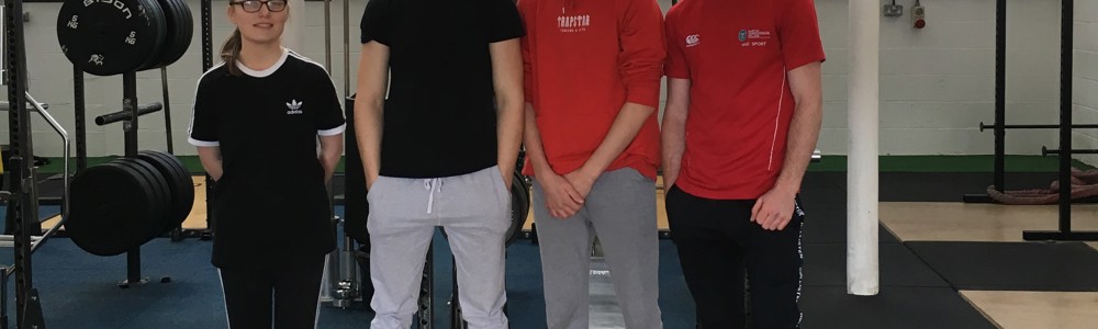 4 male and female students stood smiling on a black and wooden weightlifting platform in a gym.