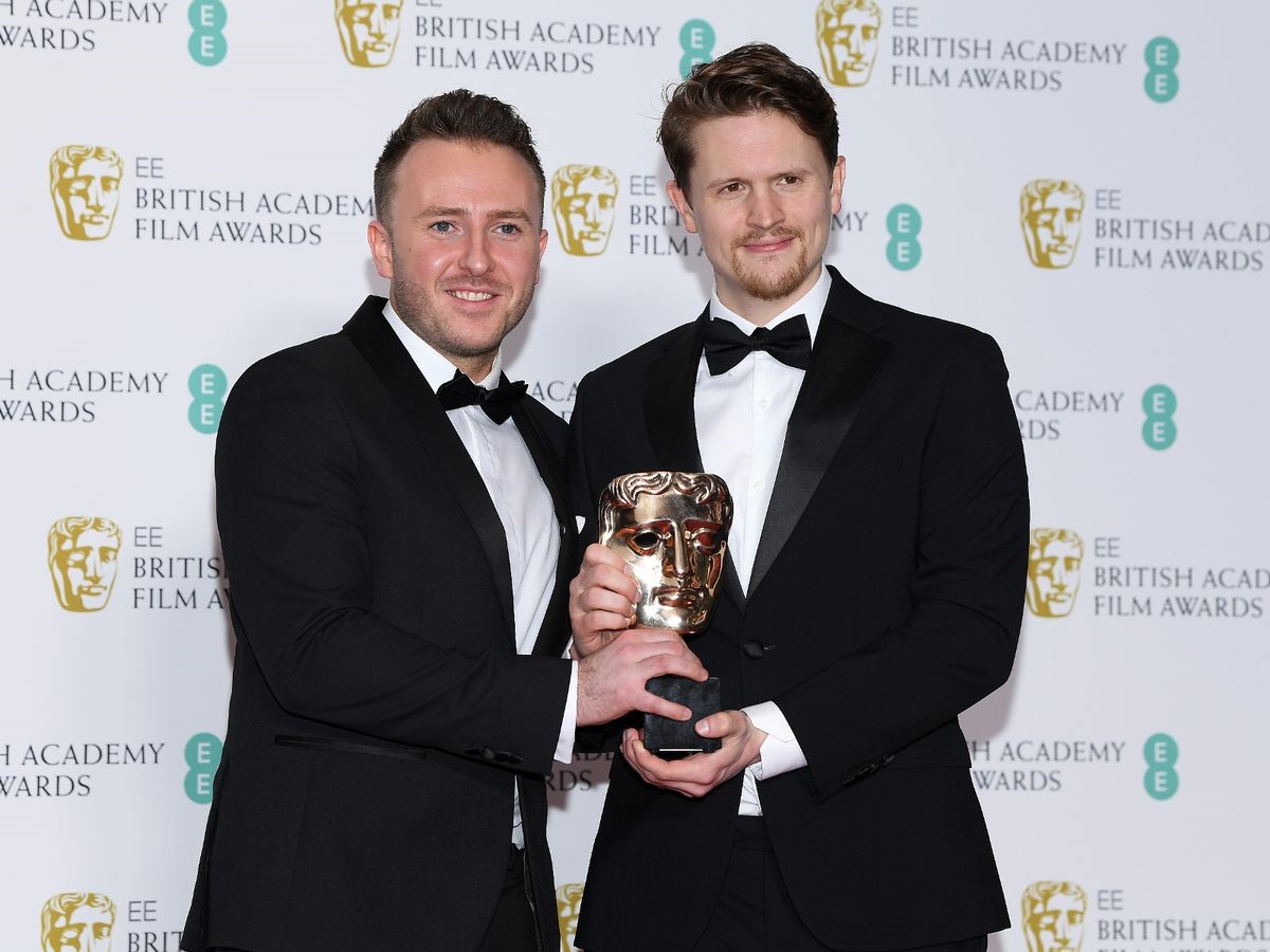 2 male adults in black tuxedos holding a Bafta award together while stood in front of a backdrop with Bafta logos on.