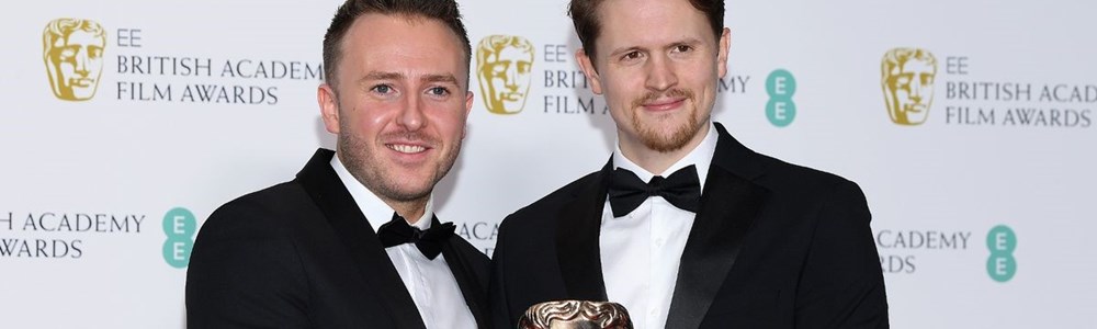 2 male adults in black tuxedos holding a Bafta award together while stood in front of a backdrop with Bafta logos on.