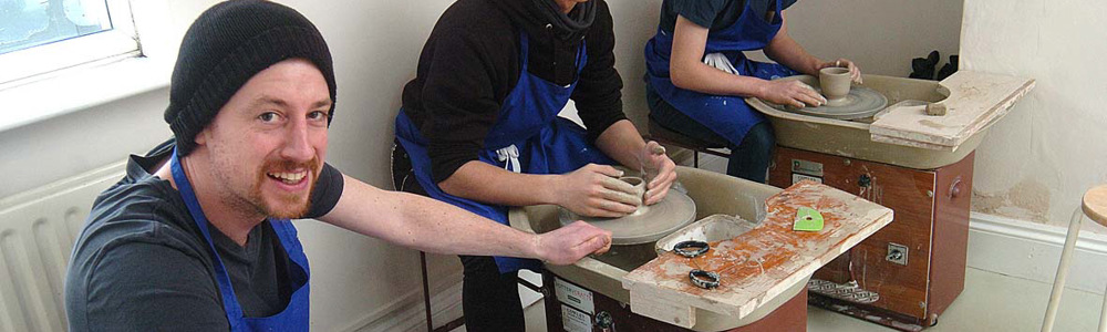 3 smiling male students in blue aprons molding clay pottery.