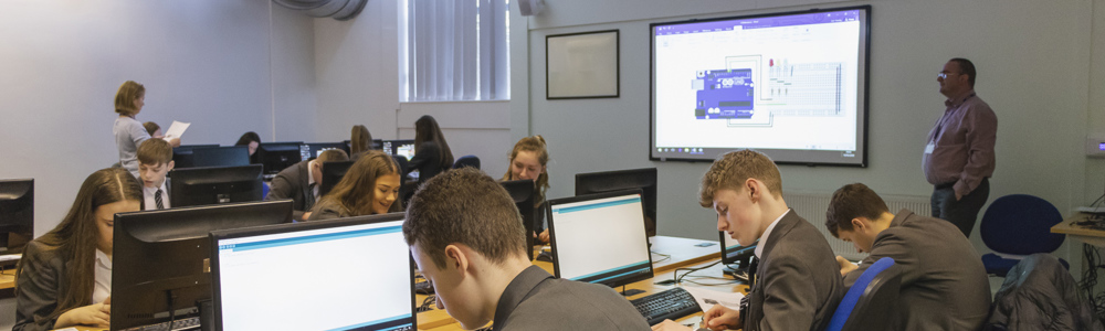 Class of students all in business attire and sat working at computers, whilst a lecturer stands at a whiteboard.