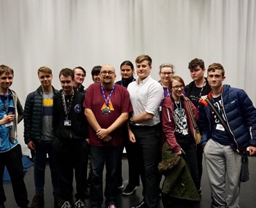 Large group of computing students stood smiling in front of white drapes