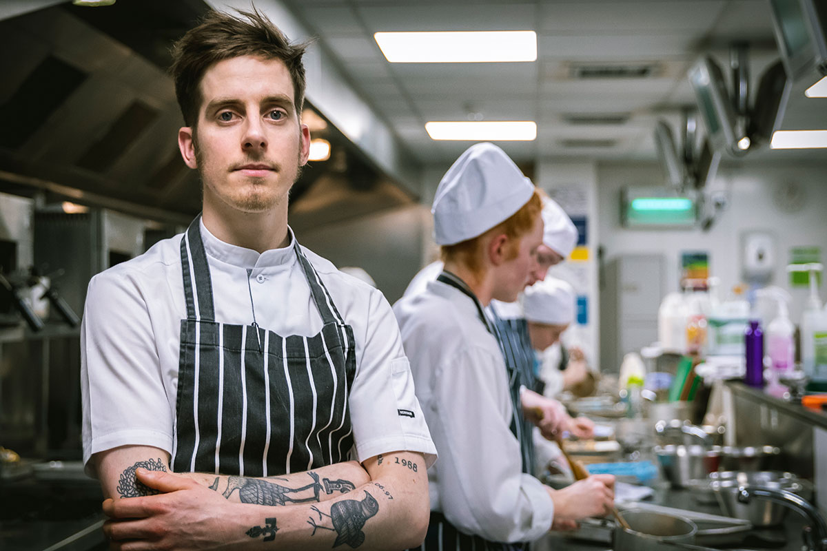 Male chef stood in commercial kitchen whilst wearing a navy striped apron with his arms crossed over his chest. Students in chef uniforms cooking in the background.