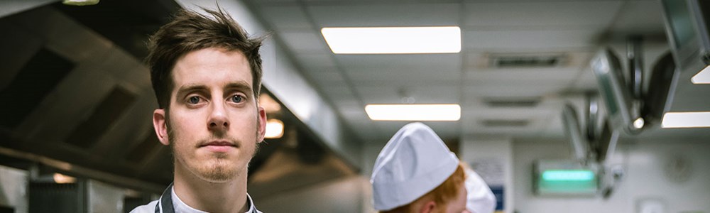 Male chef stood in commercial kitchen whilst wearing a navy striped apron with his arms crossed over his chest. Students in chef uniforms cooking in the background.