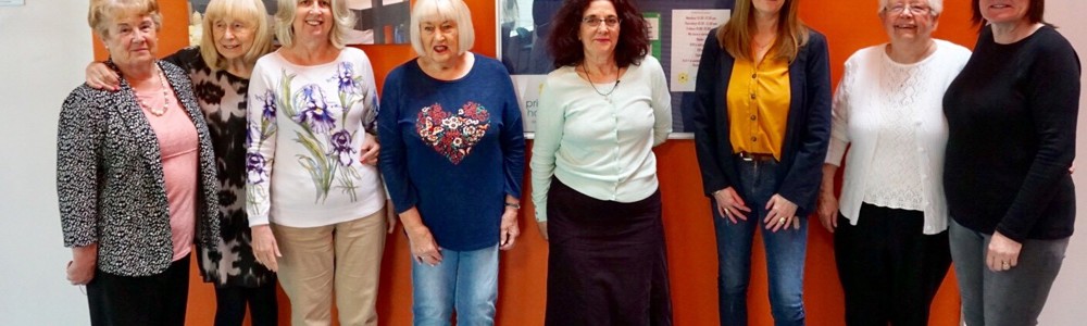 Group of female adults of varying ages stood smiling in front of orange wall