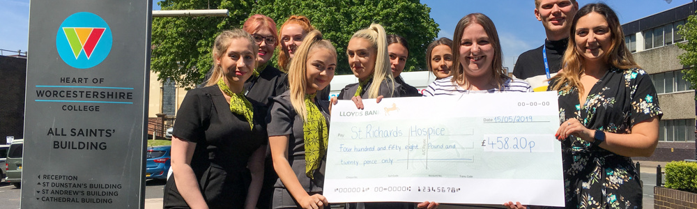 Group of students wearing travel and tourism uniforms stood outside and smiling next to HoW College sign whilst holding a giant cheque.