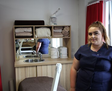 Female student wearing navy beauty therapy uniform and stood at a beauty therapist station with a massage bed.