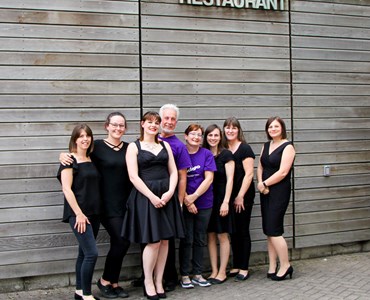Group of adults dressed smartly in black and purple. Stood smiling underneath Archer's Restaurant sign outside.