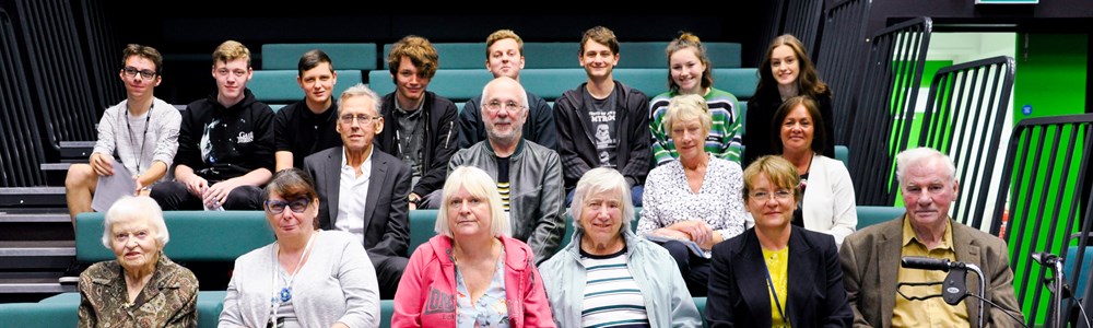 Group of senior adults and college students sat on green theatre benches in a black TV studio