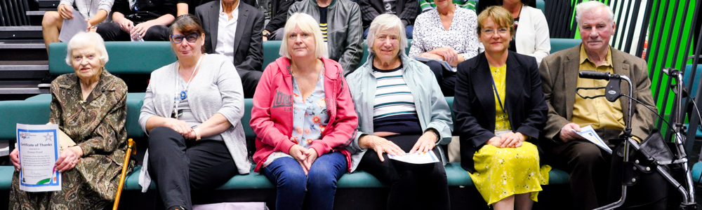 Group of senior adults and college students sat on green theatre benches in a black TV studio