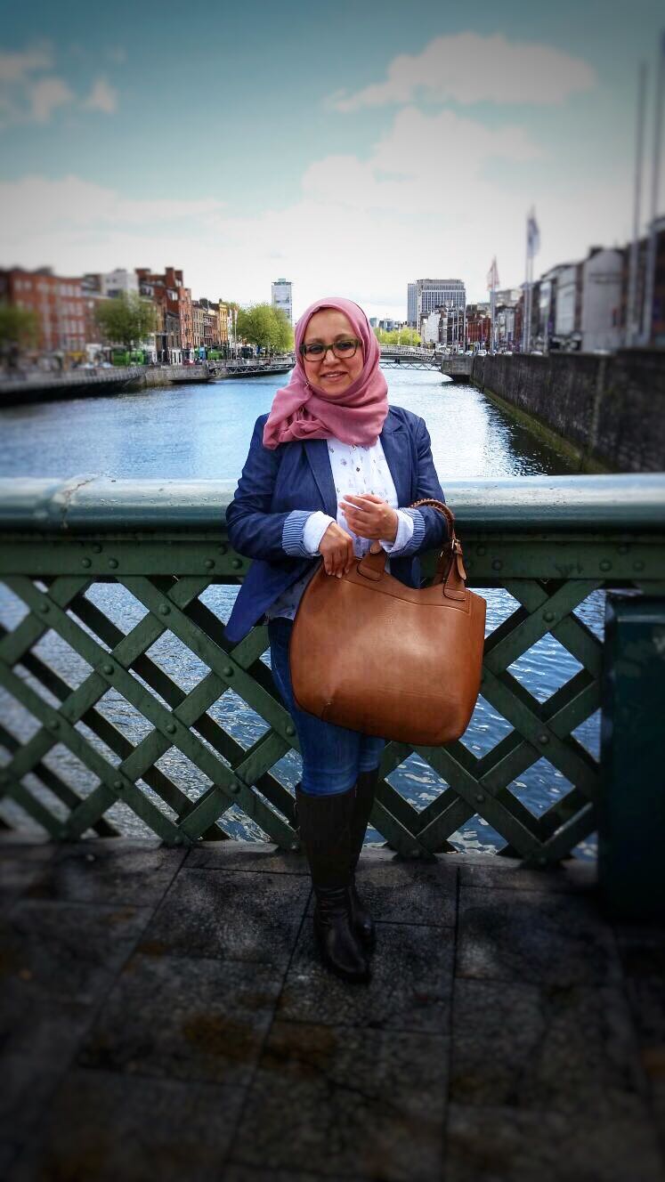 Female wearing a pink hijab and navy blazer stood holding a large brown leather handbag in front of a green bridge. Water and city can be seen in the background.