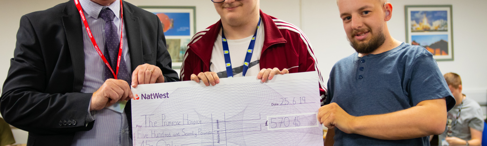 Male lecturer in grey suit stood holding a giant cheque with 2 male students.