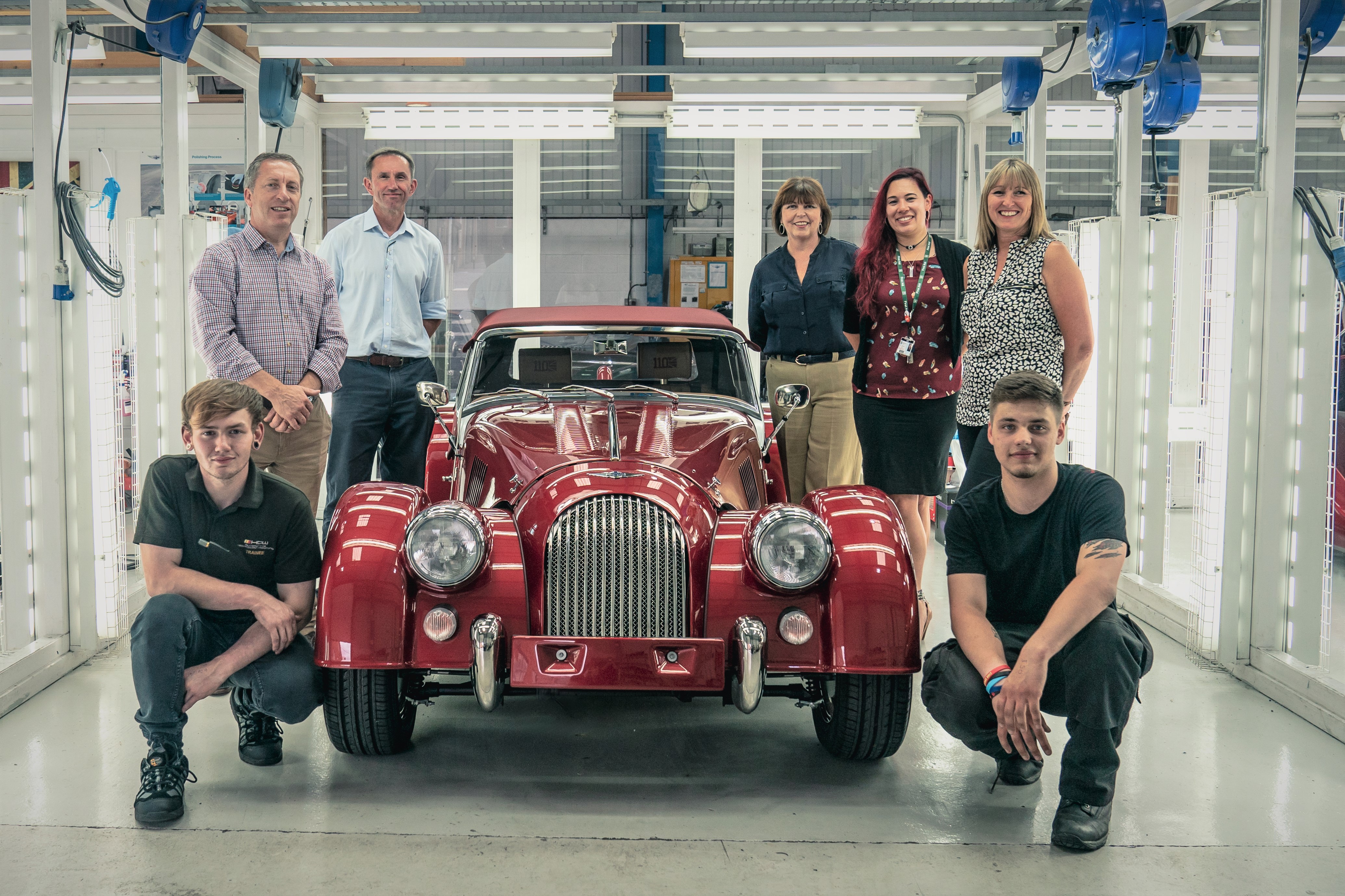 Males and females stood around a cherry red vintage car in a brightly lit white garage