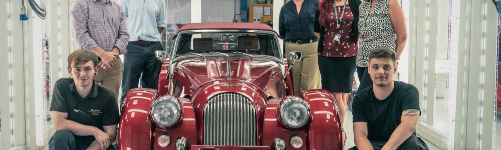 Males and females stood around a cherry red vintage car in a brightly lit white garage