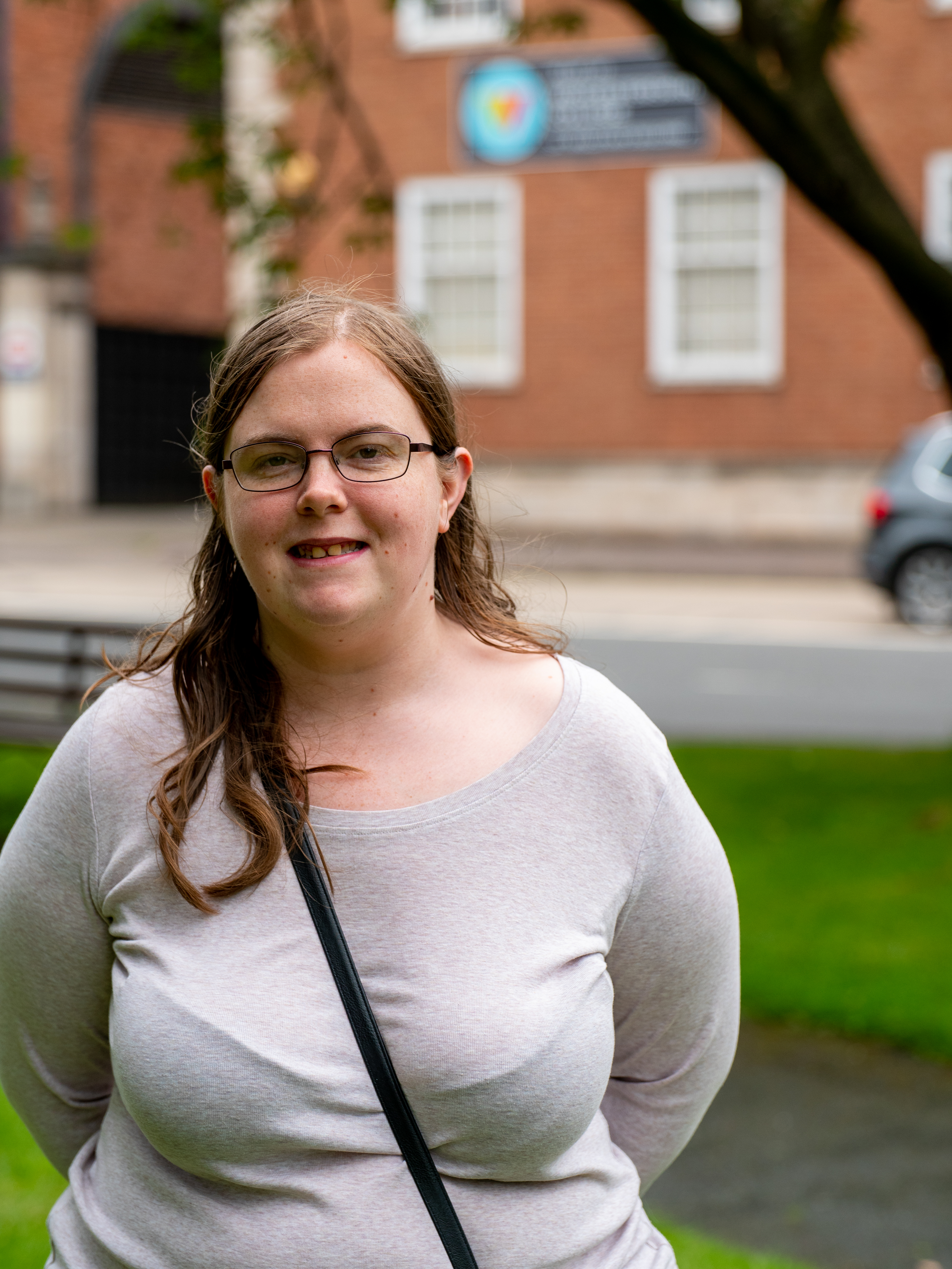 Smiling image of female in white shirt with black cross body handbag. HoW College logo and be seen in the background on a brick building.