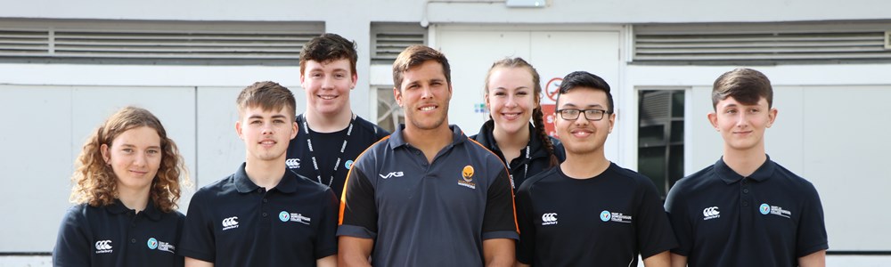 Group of 6 smiling sports students wearing matching college polo tops. Male adult smiling in the centre of the group and wearing a rugby shirt with the Worcester Warriors logo on.